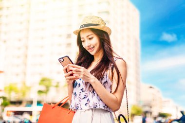 Genç ve çekici Asyalı kadın alışveriş torbaları tutuyor. Sokakta cep telefonu izliyorlar.