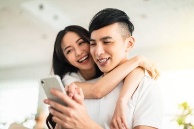  Evdeki oturma odasında cep telefonuna bakarken kucaklaşan mutlu genç çift.