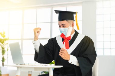 Tıp maskesi takıp, dizüstü bilgisayarı izleyerek, diplomayı tutarak ve başarıyı kutlayarak mutlu mezuniyet. 