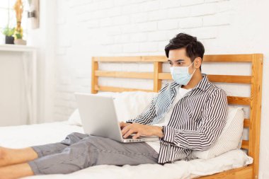  Genç adam tıbbi maske takıyor, yatakta yatıyor, çalışmak için dizüstü bilgisayar kullanıyor.