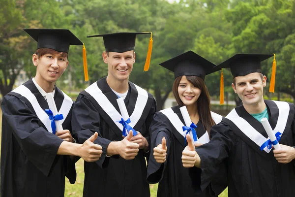 Happy students in graduation gowns showing diplomas with thumbs — Stock Photo, Image