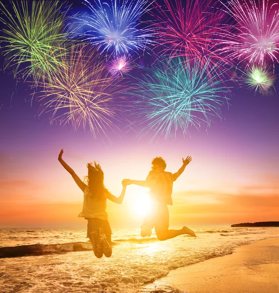 Young couple jumping on the beach with fireworks background — Zdjęcie stockowe