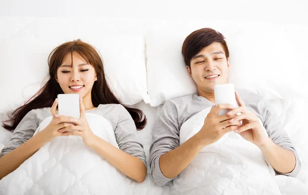 Feliz pareja joven en una cama con teléfonos inteligentes — Foto de Stock