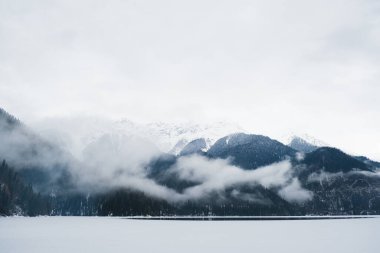 Karlı dağların ve alçak bulutların arka planında buzla kaplı kış gölü. Dağ manzarası