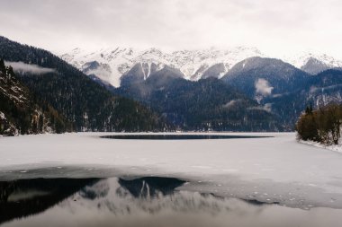 Karlı dağların ve alçak bulutların arka planında buzla kaplı kış gölü. Dağ manzarası