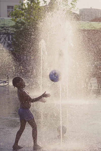 Gamin jouer avec une balle dans une fontaine d'eau ensoleillée — Photo