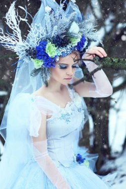 Mavi elbiseli güzel bir kadın portresi ve kış ormanlarında yürüyen bir çiçek tacı. Prenses gibi giyinmiş.