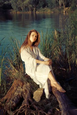 Güzel kız İvana Kupala 'yı kutluyor geleneksel beyaz elbiseli gün batımında göl kenarında oturuyor.