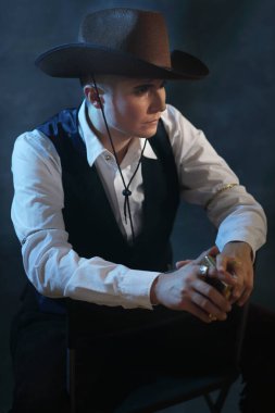 Klasik kovboy kıyafeti giymiş çift cinsiyetli bir kadın modelin yan görünüş portresi. Güçlü erkeksi estetik, düşünceli ifade, karamsar stüdyo ışıkları mavi sis ve dumanla. Cinsiyet akışkan moda ve hikaye anlatımı için kavramsal görüntü