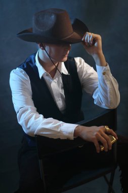 Klasik kovboy şapkası ve kıyafeti içinde erkek bir kadın modelin sinematik yakın çekimi. Şapka kaplayan gözlerle gizemli bir poz, kenarı ayarlamak. Moody stüdyo arka planında mavi ışık ve duman var. Gizemli romanlar, cinsiyet çeşitliliği ve...