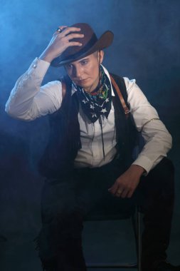 Vahşi batı kanun kaçağı gibi giyinmiş, erkeksi bir kadın modelin yüksek kaliteli stüdyo portresi. Mavi ışık ve puslu kasvetli bir atmosfer. Cinsiyet çeşitliliği, karakter tasarımı ve kitap kapakları için kavramsal resim