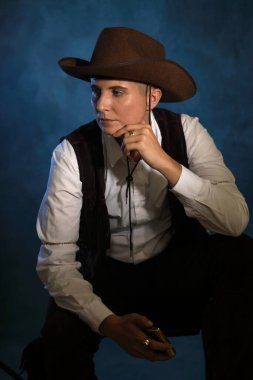 Klasik kovboy şapkası ve kıyafeti giymiş düşünceli çift cinsiyetli bir kadın. Mavi ve sıcak ışıklandırmalı, çenesinde bir el olan karamsar sinema stüdyosu portresi. Karakter tasarımı, erkeklik kavramları ve hikaye anlatımı için ideal..