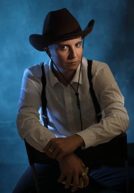 Kendinden emin çift cinsiyetli kadın model doğrudan kameraya bakıyor. Eski kovboy kıyafeti giymişti. Huysuz bir stüdyo arkaplanı, mavi ışığı ve dumanı vardı. Güçlendirme, çeşitlilik ve karakter anlatımı temaları için mükemmel