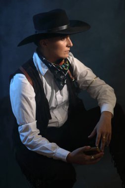 Vahşi batı karakteri gibi giyinmiş maskülen bir kadın modelin yan görünümü. Moody stüdyo atmosferi, mavi ışık, duman ve klasik giysiler. Hikaye anlatma, cinsiyet sıvısı kavramları ve kitap kapağı tasarımı için mükemmel.