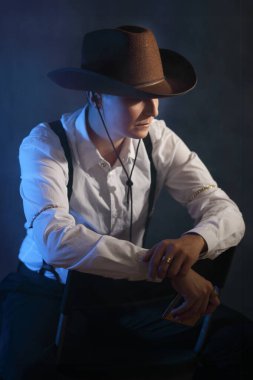 Vahşi batı kovboyu gibi giyinmiş düşünceli bir çift cinsiyetli kadın modelin portresi. Mavi ışıklandırmalı ve dumanlı karamsar stüdyo atmosferi. Güç, çeşitlilik ve sinematik hikaye anlatımı için kavramsal görüntü