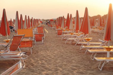 İtalya 'nın Bibione kentinin kumlu sahilinde altın bir günbatımında turuncu plaj şemsiyeleri ve şezlonglar. Yaz tatili ve seyahat temaları için mükemmel.