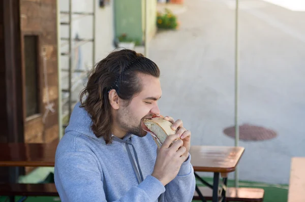 Young guy with long hair eats big sandwich — Stock Photo, Image