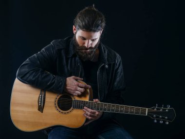 Genç sakallı adam siyah arka planda oturup akustik gitar çalıyor..
