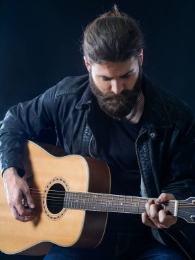Genç sakallı adam siyah arka planda oturup akustik gitar çalıyor..