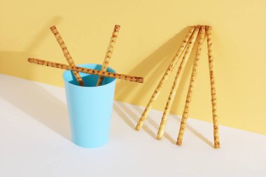 Mavi plastik bardakta susam tohumlu ekmek çubukları ve beyaz-sarı arka planda