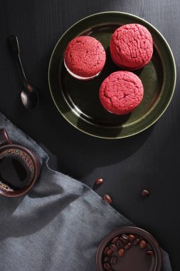 Kırmızı kadife sandviç kurabiyeleri ve siyah ahşap masada bir fincan kahve.