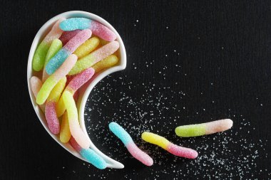 Sour fruit gummy candies in the form of worms in bowl on black stone background, top view