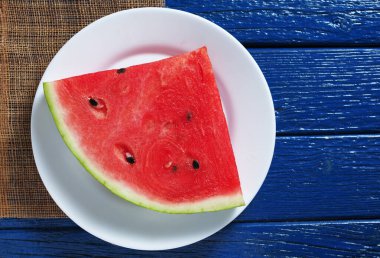 Mavi ahşap masada beyaz bir tabakta sulu bir karpuz dilimi.