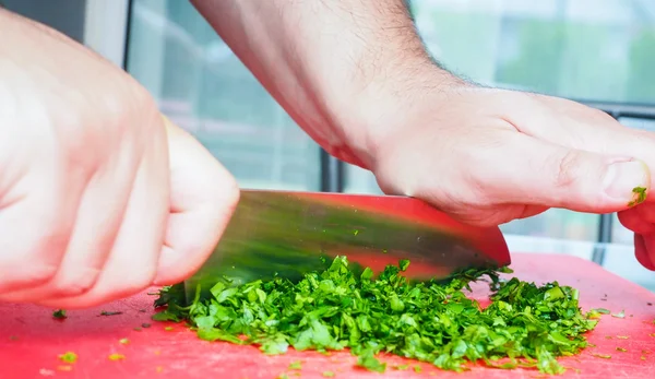 Chef masculino cortando perejil con cuchillo grande en tabla de cortar roja — Foto de Stock