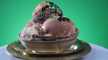 Gümüş bardakta çikolatalı dondurma topları, tatlı serpiştirilmiş, parlak renkli bir arka planda yavaşça dönüyor..