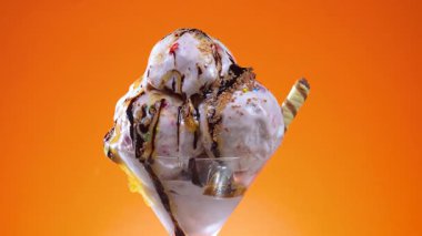 Bir cam kasede vanilyalı dondurma, çikolata serpilmiş ve şurup serpiştirilmiş, parlak renkli bir arka planda yavaşça dönüyor..