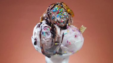 Meyve şurubuyla ıslatılmış cam kasede çikolatalı dondurma topları ve parlak renkli bir arka planda yavaşça dönüyor..