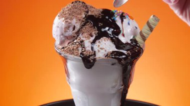 Bir cam kasede vanilyalı dondurma, çikolata serpilmiş ve şurup serpiştirilmiş, parlak renkli bir arka planda yavaşça dönüyor..