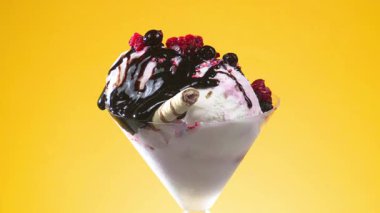 Bir cam kasede vanilyalı dondurma, çikolata serpilmiş ve şurup serpiştirilmiş, parlak renkli bir arka planda yavaşça dönüyor..