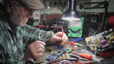 Yaşlı bir teknisyen, ölçüm aletleri ve teknik aletlerle atölyede bilgisayar donanımı tamir ediyor. Yavaş çekim.