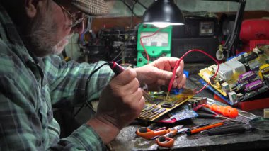 Yaşlı bir teknisyen, ölçüm aletleri ve teknik aletlerle atölyede bilgisayar donanımı tamir ediyor. Yavaş çekim.
