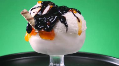 Çikolata jölesi serpilmiş cam bir kasede meyveli dondurma. Parlak bir arka planda yavaşça dönüyor..