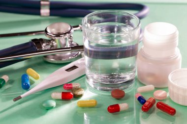 Bir bardak su, bir termometre, haplar ve masanın üstünde renkli bir arka planda steteskop. Yakın plan..