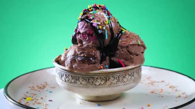 Metal bir kasede çikolatalı dondurma, yenilebilir konfeti serpiştirilmiş, parlak bir arka planda yavaşça dönüyor..