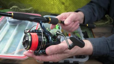 Stüdyo kompozisyonunda bir masada spor balıkçılık ekipmanları, bir ağın arka planına karşı. Balıkçının elleri, olta takımını kontrol eder..