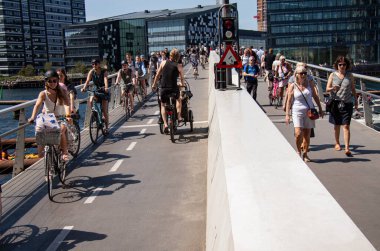 Birçok insan güneşli bir havada limanın yanında bisiklet sürüyor ve modern bir yaya ve bisiklet köprüsünde yürüyor. Kopenhag, Danimarka - 5 Haziran 2021. 