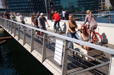 Birçok insan güneşli bir havada limanın yanında bisiklet sürüyor ve modern bir yaya ve bisiklet köprüsünde yürüyor. Kopenhag, Danimarka - 5 Haziran 2021. 