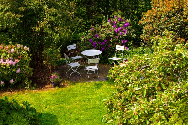 Yeşil çimenli güzel bir bahçe manzarası ve yapraklı renkli çiçek yatağı ve gölgede beyaz bahçe mobilyaları. Yuvarlak masa ve sandalyeler..