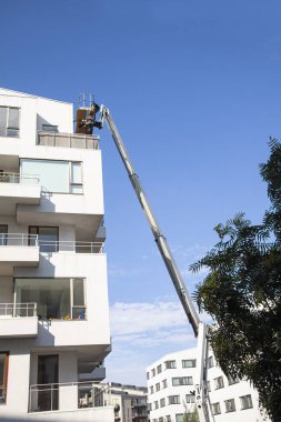 Yükseklerde eklemli bir asansörde çalışan bakım işçileri, konut binasının ön cephesini tamir ediyorlar. Mavi gökyüzü arka planı. Sanayi iş imajı.