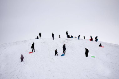 Siluetteki insanlar karlı bir tepede yetişkinler ve çocuklar kızakla kayarken eğleniyorlar. Kış eğlencesi.