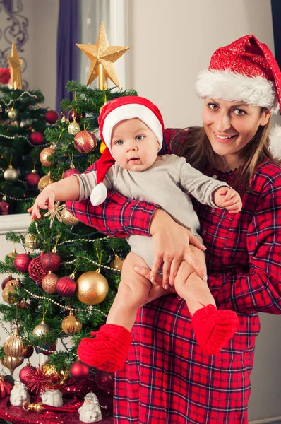 母と彼女の美しいです赤ちゃん男の子Posing横にクリスマスツリーとともにサンタクラスの帽子 — ストック写真