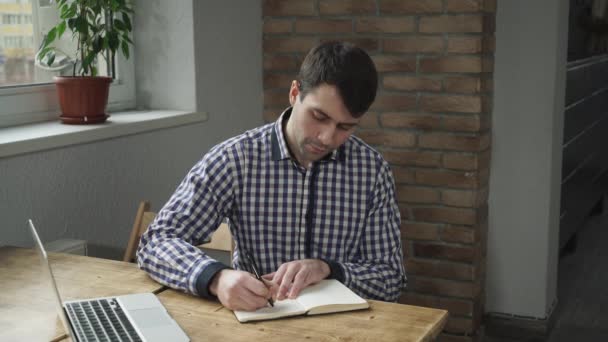 Hombre atractivo en una camisa haciendo notas en un diario y mirando un portátil . — Vídeos de Stock