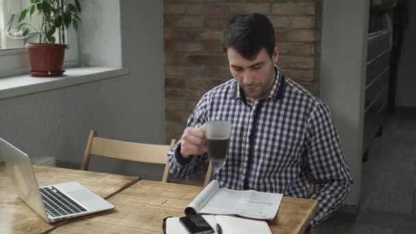 Hombre atractivo bebiendo café y leyendo el periódico, está en la mesa teléfono y un ordenador portátil . — Vídeos de Stock