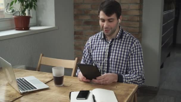 El hombre se sienta en una mesa con una tableta, auriculares en los oídos, hablando en línea . — Vídeos de Stock