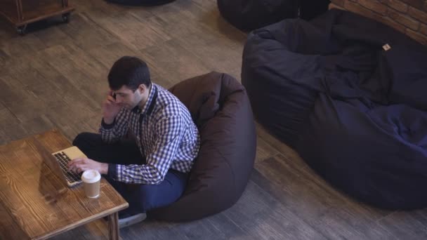 El gerente conduce el teléfono, mirando a la computadora que está sobre la mesa, de pie junto a un vaso de café . — Vídeo de stock