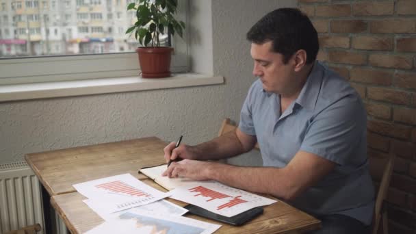 Gerente sentado na mesa, tomando notas em um caderno, em uma mesa papéis espalhados . — Vídeo de Stock